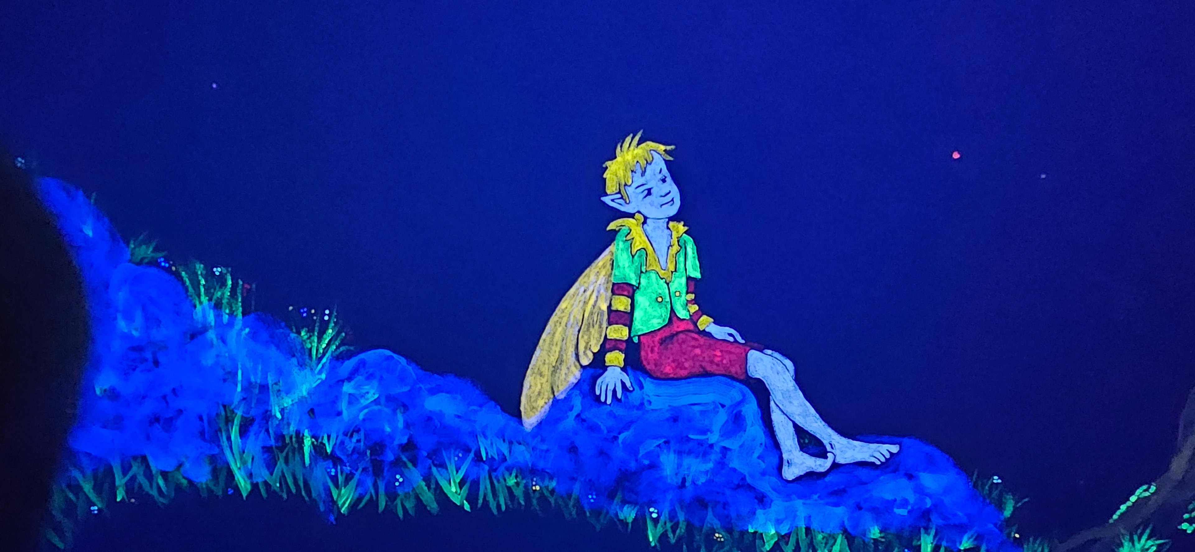 Glowing fairy boy with yellow hair sitting on a glowing blue mossy rock under black light in the Fantasy Room